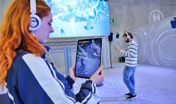 A visitor with headphones holds an iPad showing her augmented-reality flower, while another visitor sings in the space